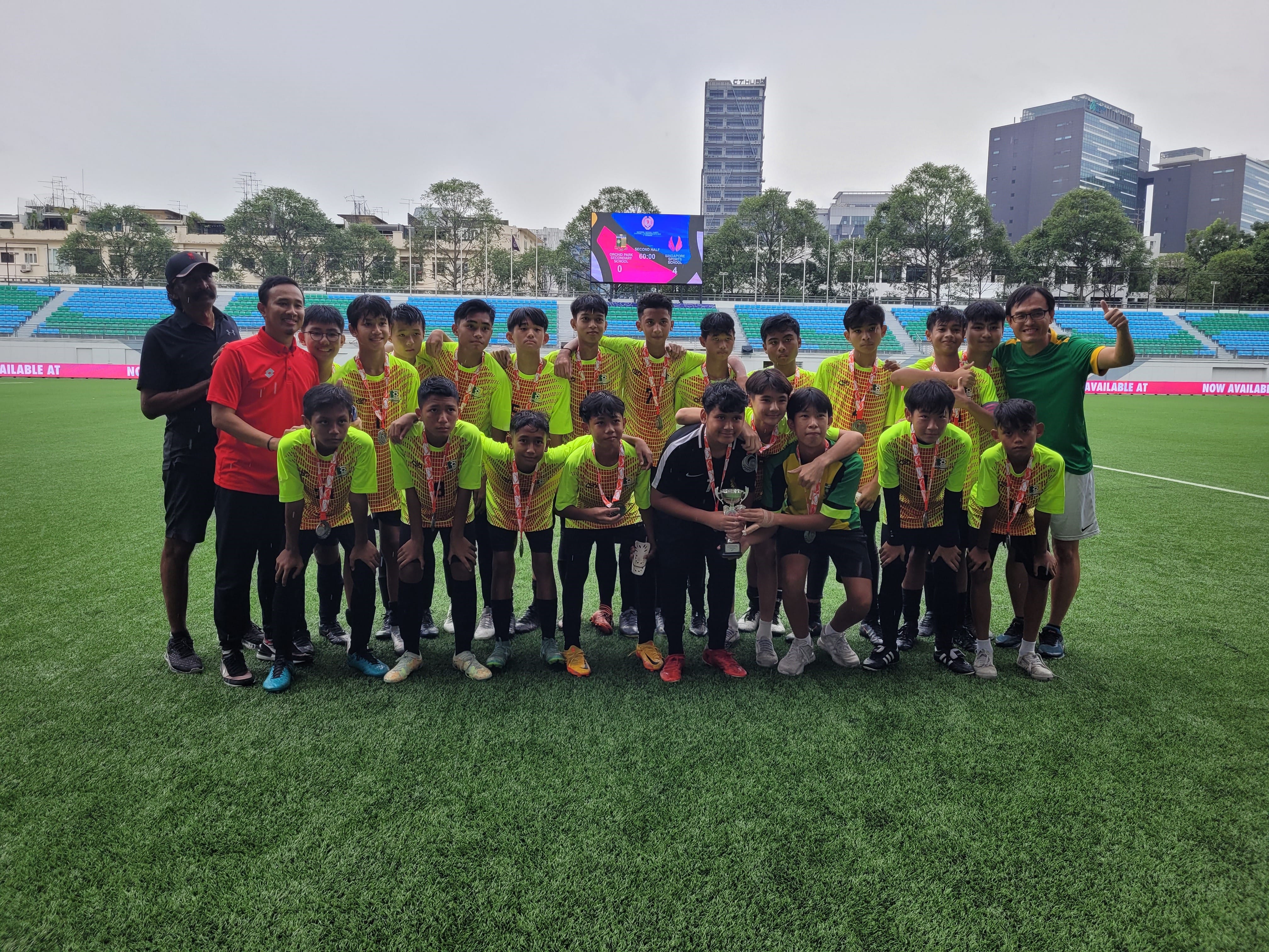 A group photo of a C-Division youth football team and their coaches celebrating on a large stadium pitch. The players are wearing neon yellow patterned jerseys with silver runner-up medals around their necks, proudly holding up a trophy in front of stadium seating and a city skyline.